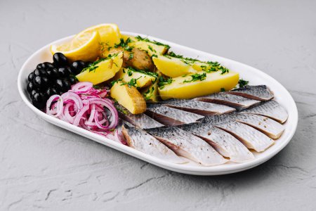 Marinated herring on a plate with potatoes, pickled onions, olives, and lemon slices