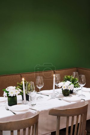 Photo for Cozy dining table setup with lit candles, fresh flowers, and elegant glassware against a green backdrop - Royalty Free Image