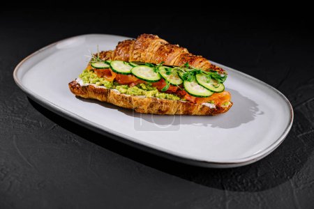 Téléchargez les photos : Sandwich croissant élégant avec saumon fumé, avocat, concombre et herbes fraîches sur une assiette blanche - en image libre de droit
