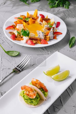 Vertical image showcasing a colorful salad with cheese and strawberries and a shrimp appetizer with lime