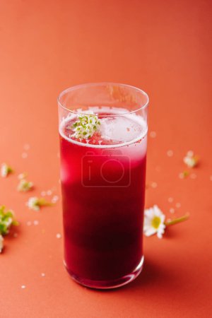 Vibrant red berry smoothie in a tall glass, decorated with small white flowers on an orange backdrop