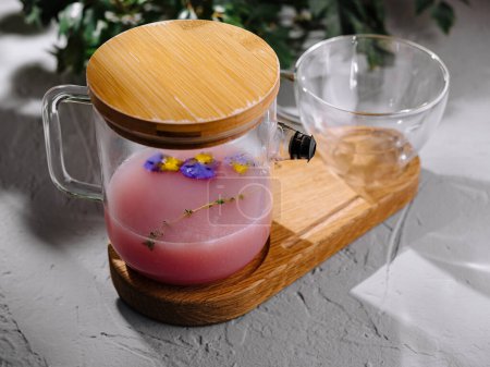 Tetera de vidrio transparente con té de hierbas rosa y flores comestibles en una bandeja de bambú, estilo sobre un fondo de hormigón