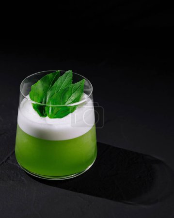 Elegant green cocktail with a frothy top and fresh basil garnish on a dark backdrop