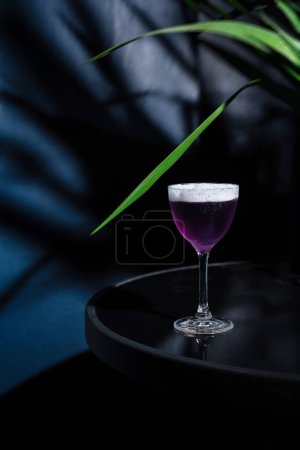 Stylish purple cocktail in a classic glass, set against a moody, dark backdrop with green plant accents