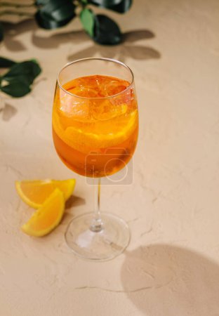 Glass of vibrant aperol spritz on a sandy surface accompanied by fresh orange wedges
