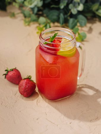 Iced strawberry lemonade garnished with fresh mint and lemon in a mason jar on a sandy background