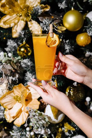 Primer plano de una mano sosteniendo un vaso de bebida caliente de vacaciones con decoraciones navideñas en el fondo
