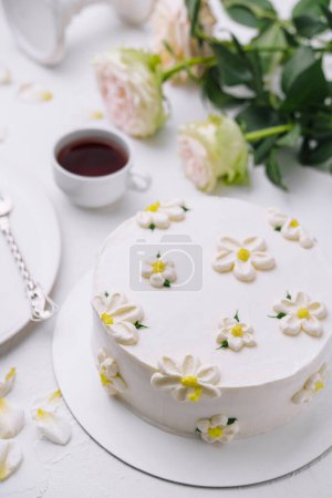 White frosted cake adorned with flowers, alongside tea and roses, on a chic table setting