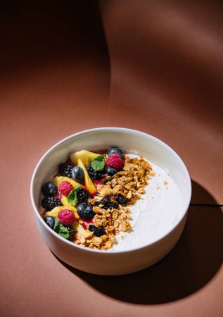 Healthy bowl of yogurt with granola and mixed fruits, presented on a stylish brown backdrop with shadows