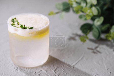 Close-up of a lemon cocktail topped with foam and a sprig of thyme, on a textured surface
