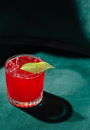Vibrant red cocktail with ice and a lime leaf, presented on a luxurious green velvet backdrop