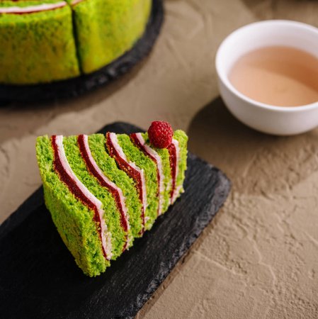 Rebanada de vibrante pastel de té verde matcha, decorado con frambuesas, junto a un pastel completo y una taza de té