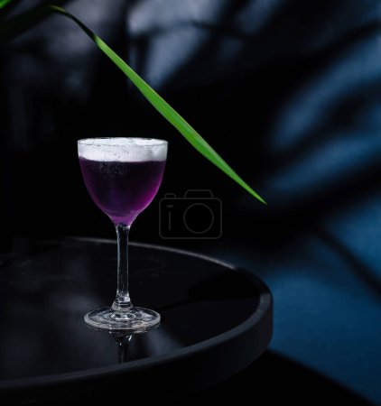 Cocktail violet élégant dans un verre classique, sur fond sombre et humide avec des accents végétaux verts