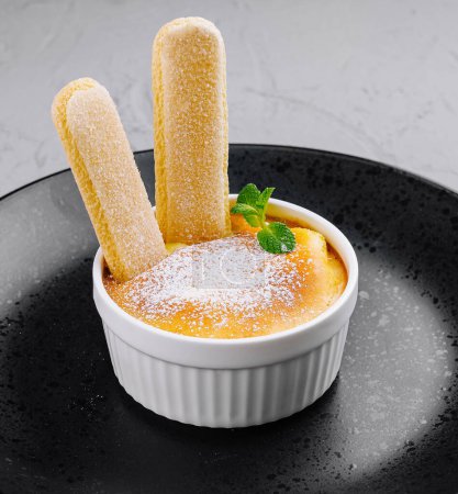 Téléchargez les photos : Crème brulée classique dans un ramequin blanc aux doigts de dame, saupoudré de sucre en poudre sur une assiette sombre - en image libre de droit