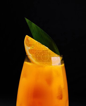 Vibrant orange drink in a glass, garnished with a slice and leaf, against a dark backdrop