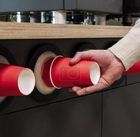 Nahaufnahme einer Hand, die in einer Cafeteria einen roten Pappbecher aus einem modernen Becherspender zieht