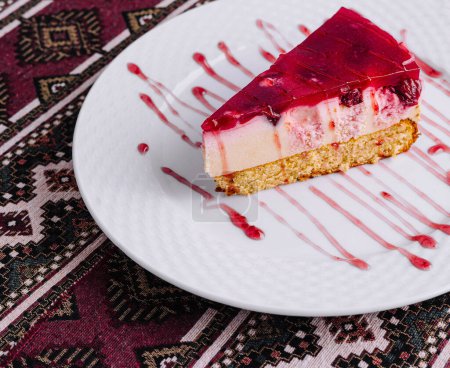 Tempting slice of raspberry cheesecake with syrup on a white plate, over a patterned tablecloth