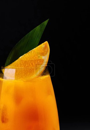 Vibrant orange drink in a glass, garnished with a slice and leaf, against a dark backdrop