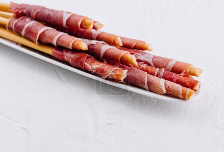 Elegante Grissini-Brotsticks in Prosciutto-Verpackung auf weißem Teller vor strukturiertem Hintergrund