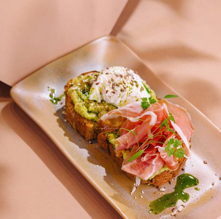 Elegant avocado toast topped with cured prosciutto, a poached egg, and fresh herbs on a stylish plate