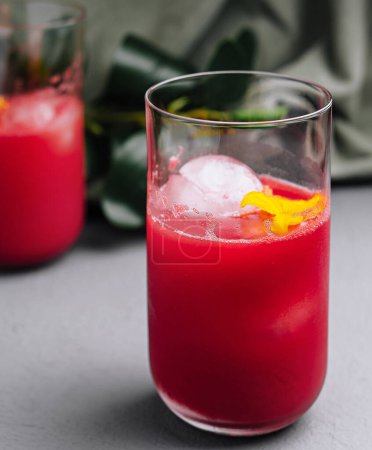 Two glasses of vibrant red citrus cocktail with ice, garnished with lemon zest