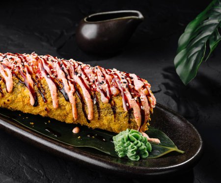 Delicious tempura sushi roll garnished with sauce, served with ginger and wasabi on a sleek black plate