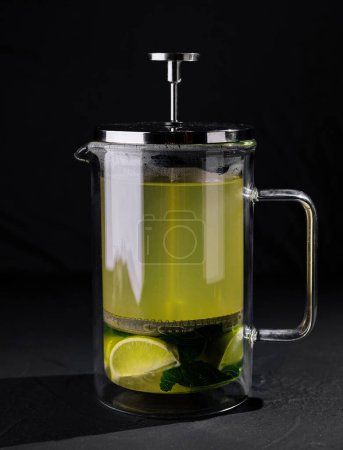 Presse française en verre transparent remplie de thé au citron vert chaud, de feuilles de menthe et de tranches de citron vert sur un fond sombre