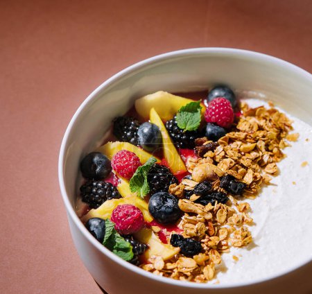 Healthy bowl of yogurt with granola and mixed fruits, presented on a stylish brown backdrop with shadows
