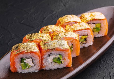 Elegant sushi rolls topped with gold leaf, served on a ceramic dish against a dark textured background