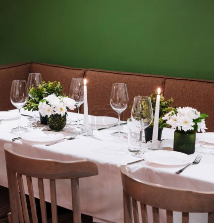 Gemütliche Esstisch-Einrichtung mit brennenden Kerzen, frischen Blumen und eleganten Gläsern vor grünem Hintergrund