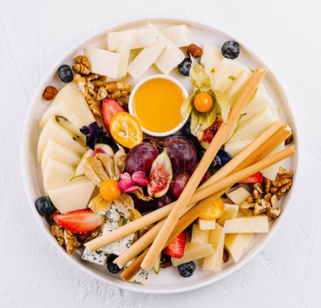 Elegante tabla de quesos con una variedad de quesos, frutas frescas, frutos secos y miel, dispuestos en una superficie blanca