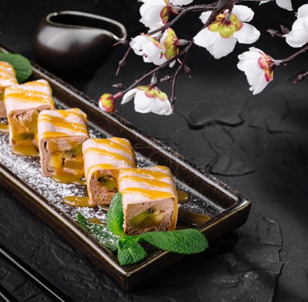 Elegant sushi rolls with kiwi and banana served on a platter with a sakura branch, on a dark stone background