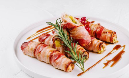Gourmet bacon-wrapped jalapenos filled with cheese, garnished with rosemary on a white plate