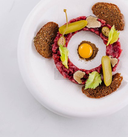 Elegant presentation of steak tartare with a quail egg, pickles, and rye bread on a white plate
