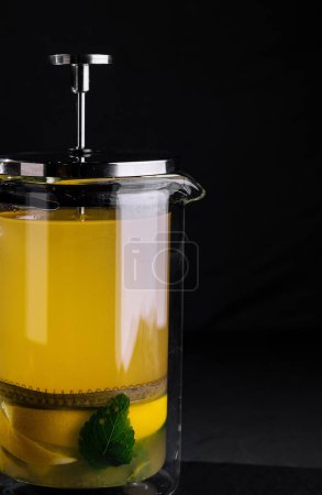 Prensa francesa de vidrio llena de té de menta caliente y limón sobre un fondo oscuro