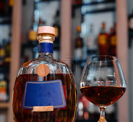Premium bottle of brandy beside a filled snifter glass presented on a vibrant bar counter with blurry shelves of bottles in the background