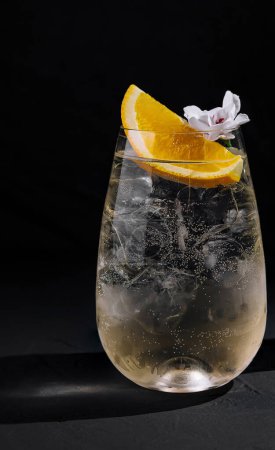 Erfrischender Gin Tonic, serviert mit Eis, Orangengarnitur und einer Blume in einem stilvollen Glas neben einer Flasche