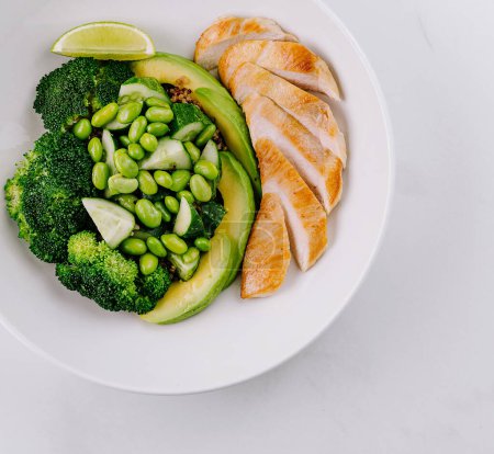 Vista aérea de una comida nutritiva con pechuga de pollo en rodajas, aguacate, edamame, brócoli y cuña de lima