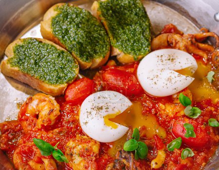 Savory shakshuka with poached eggs, pesto, and grilled bread in a skillet close up