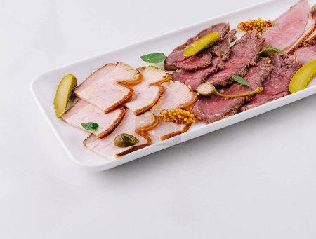 Gourmet platter of sliced deli meats served with pickles and a creamy dip on a white background