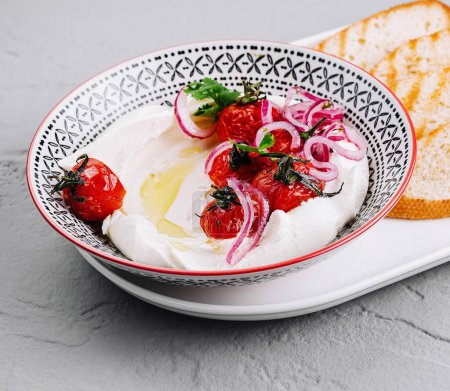 Bowl of creamy labneh garnished with herbs, served with crispy toasted bread and roasted cherry tomatoes
