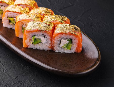 Elegant sushi rolls topped with gold leaf, served on a ceramic dish against a dark textured background
