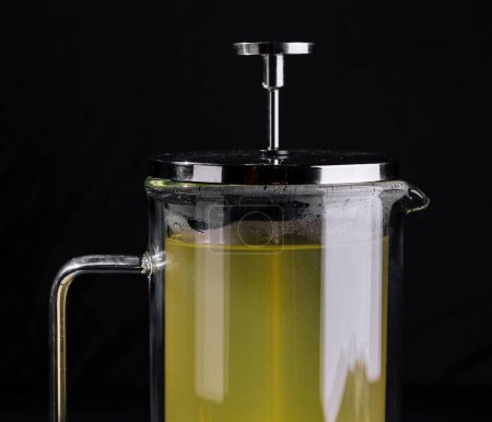 Clear glass french press filled with hot lime tea, mint leaves, and slices of lime on a dark background