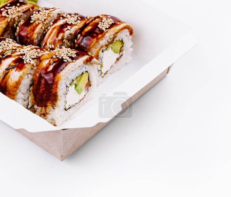Tasty eel sushi roll with avocado and sesame seeds in a paper takeout container, isolated on white