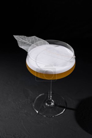 Craft cocktail in a stemmed glass with a delicate leaf adornment, showcased on a dark backdrop