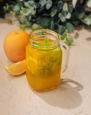 Einmachglas gefüllt mit orangefarbenem Eistee, garniert mit Minze auf einem sonnigen Tisch mit frischen Orangen