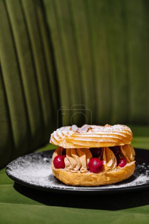 Delicious paris-brest with cream and berries on a black plate against a green background
