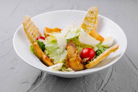 Bowl of caesar salad with grilled chicken, croutons, and cherry tomatoes on a gray background