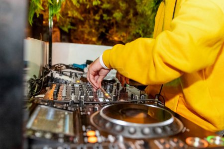 Photo for A deejay in a yellow hoodie is entertaining the crowd at a concert, skillfully mixing music on audio equipment and scratching on a mixer - Royalty Free Image