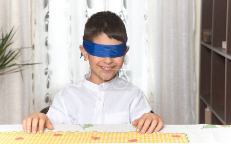 Un niño caucásico de 8 años sentado a la mesa en casa tiene los ojos vendados y sonríe.